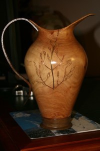 This Jug, or Ewer for those who are more sophisticated, has been turned from Golden Cyprus with brass and copper highlights. The base is Walnut. Wall thickness about 6mm. The handle, which is Jarrah, was cut roughly on the bandsaw and finished with drum sanders and anything else I could get my hands on. It is about 260mm high and was turned when the timber was green which is my preferred option. 